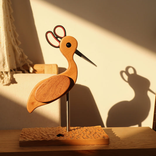 Wooden bird sculpture with scissors on a stand, casting a shadow.
