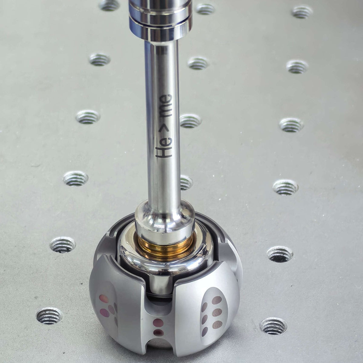 A polished metallic robotic end-effector arm with a silver cylindrical shaft and a spherical base, featuring the engraved text 'He > me' on the shaft, set against a metallic silver threaded plate.