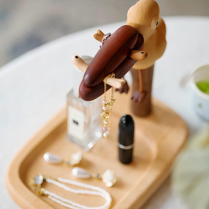 A wooden bird-shaped jewelry organizer holding a gold chain necklace, displayed on a light wood tray with other cosmetics and jewelry in a softly lit setting.