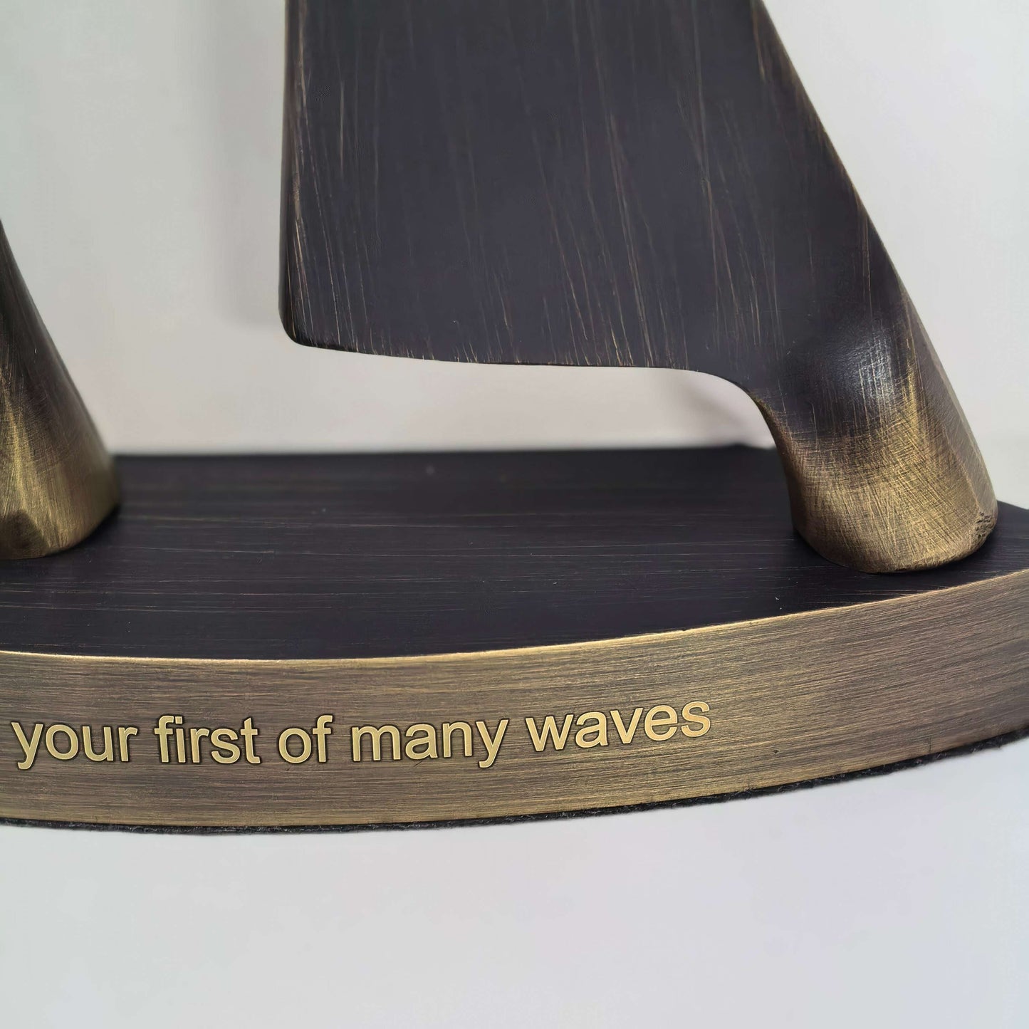 Close-up of a decorative nautical sculpture featuring dark wood-textured wave forms on a curved bronze base engraved with the phrase 'your first of many waves'.