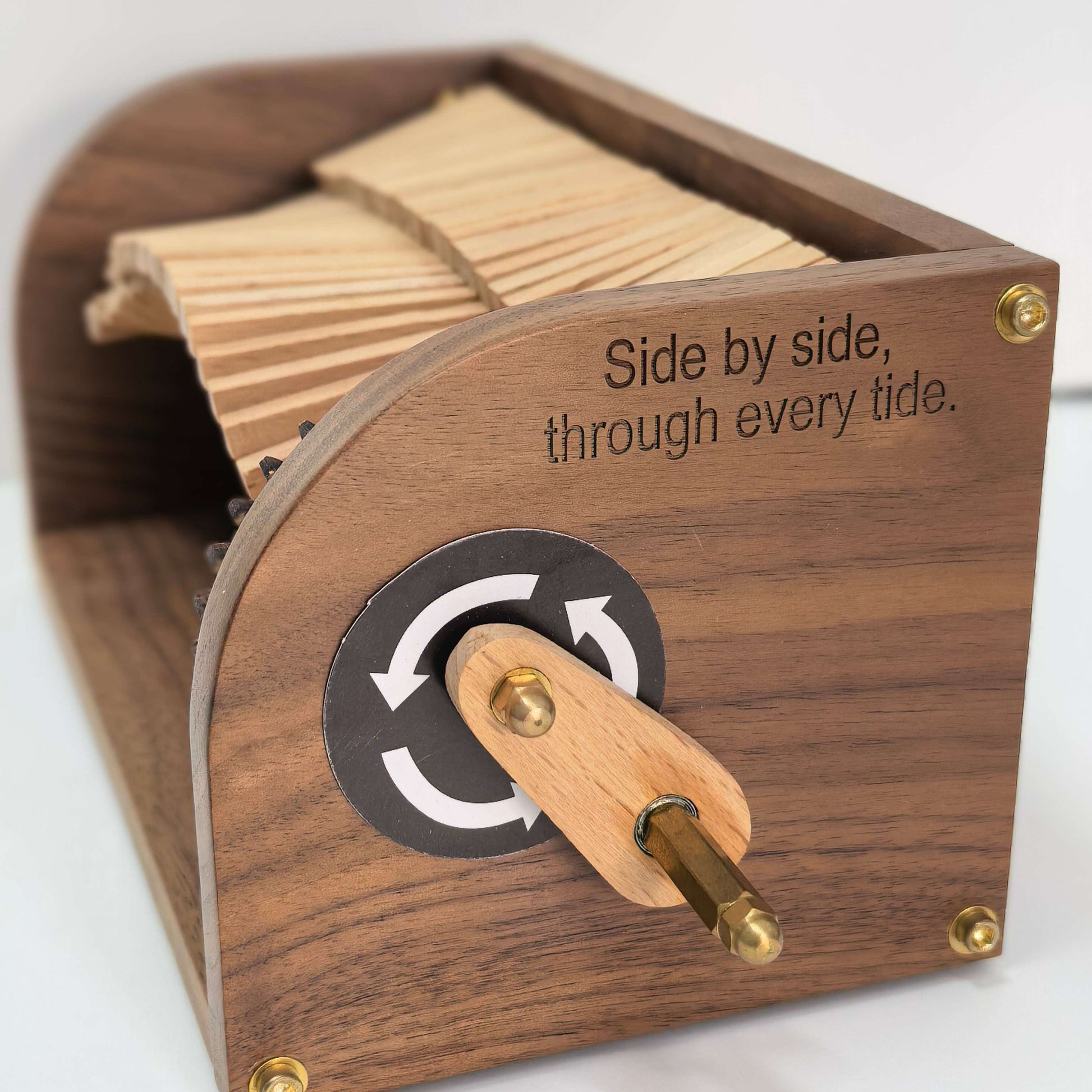 A wooden tidal clock with a dark wood body, brass screws, and a wooden crank handle. The front features an engraved black circular dial with white arrows and the text 'Side by side, through every tide.