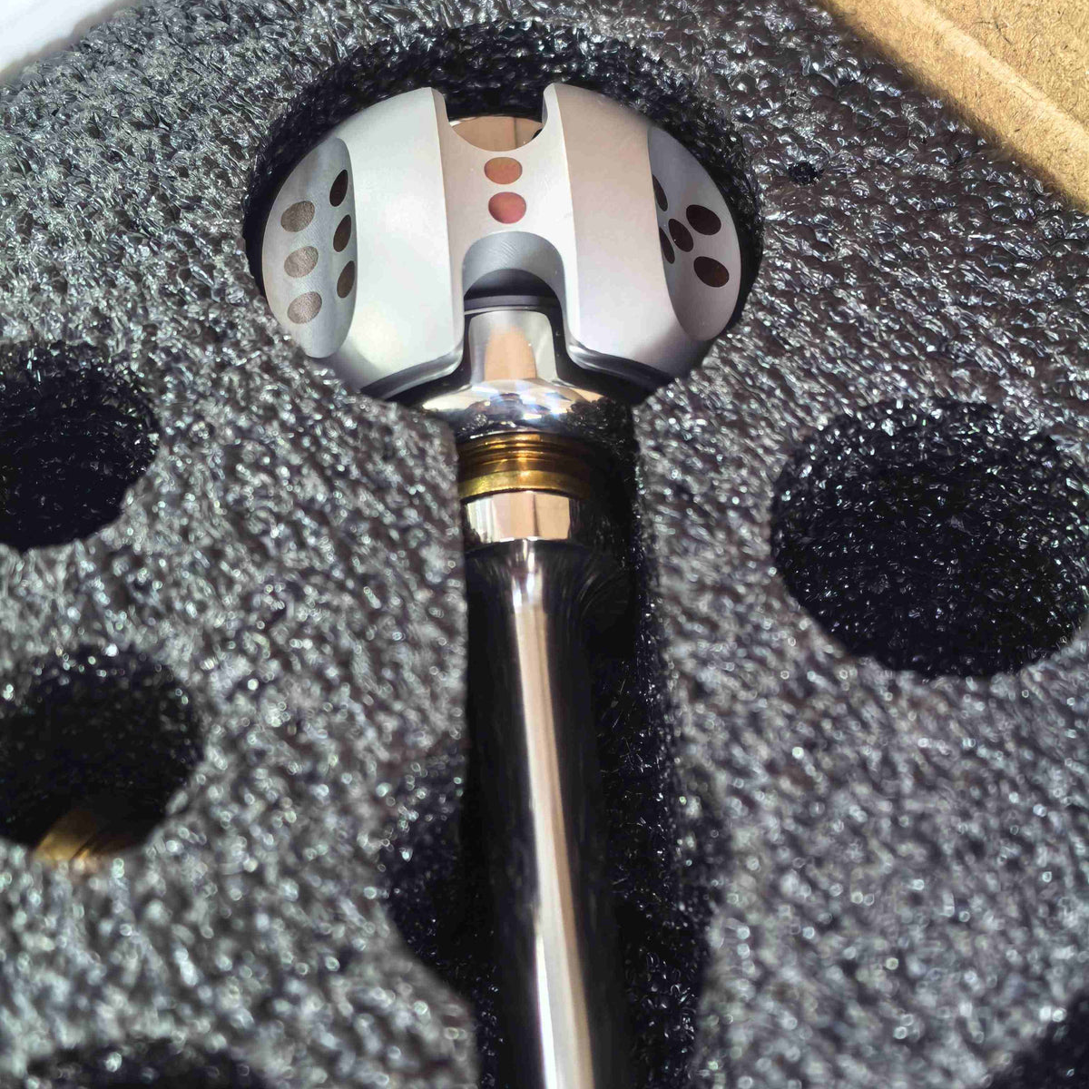 A close-up of a metallic mechanical component, possibly a sensor or valve, resting in custom black foam packaging inside a cardboard box.