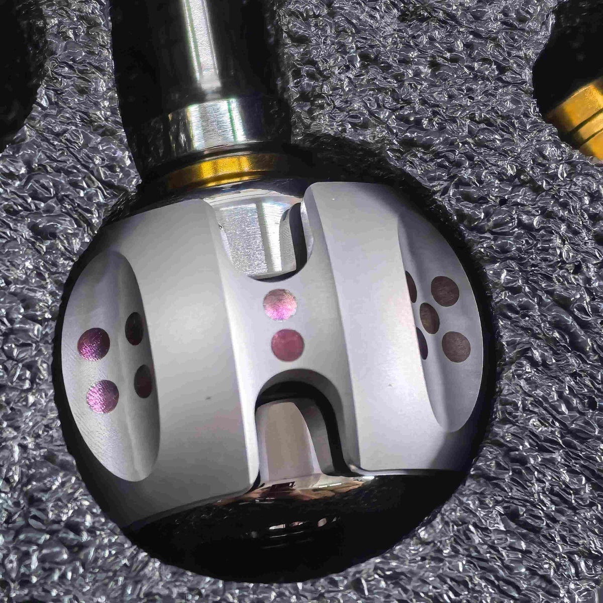 A close-up of a metallic dental drill bit head with a silver body, pink and gold accents, and a dark shaft, resting on a textured gray surface.