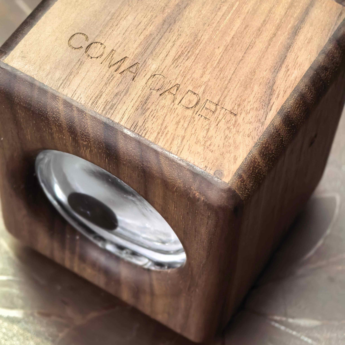A close-up of a wooden cube-shaped optical device with a circular glass lens cutout on one side and the engraved brand name 'COMA GADEY' on the top face, set against a reflective metallic surface.