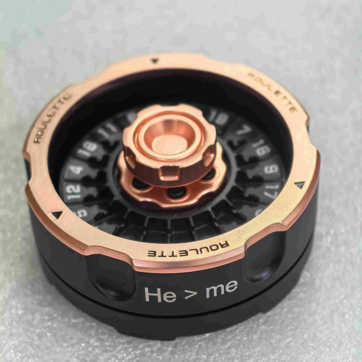 A close-up of a Roulette wheel component, featuring a black outer casing with the text 'He > me' and 'ROULETTE', a metallic rose gold outer ring, and a central copper-colored dial mechanism on a textured gray background.