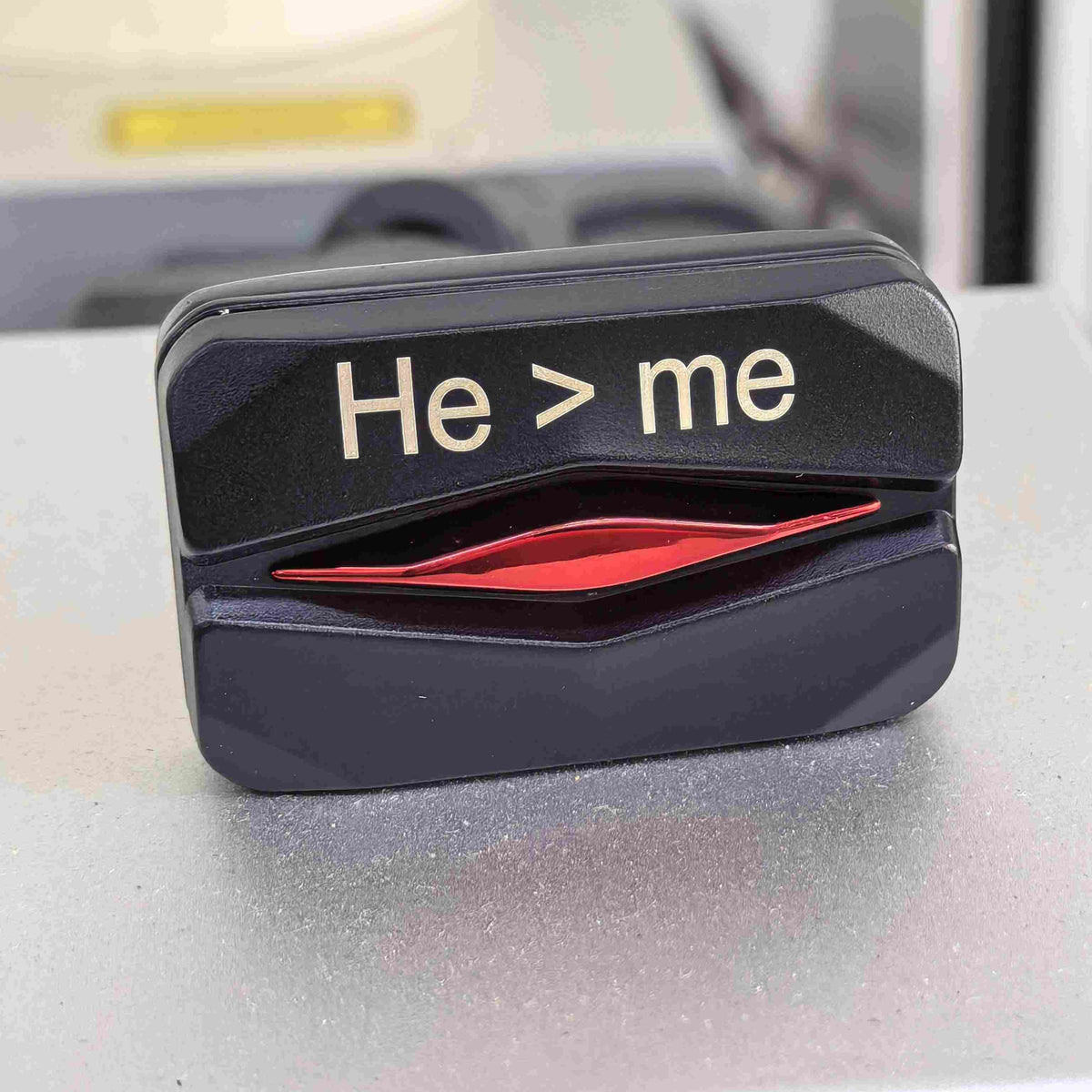 A novelty black plastic car window defroster sticker with the text 'He > me' in white, featuring a red accent graphic, shown on a car window.